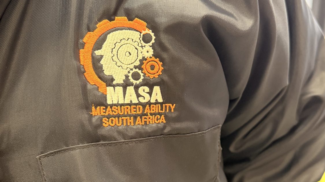 A gear and head logo with the name MASA embroidered on the chest of a grey workwear jacket