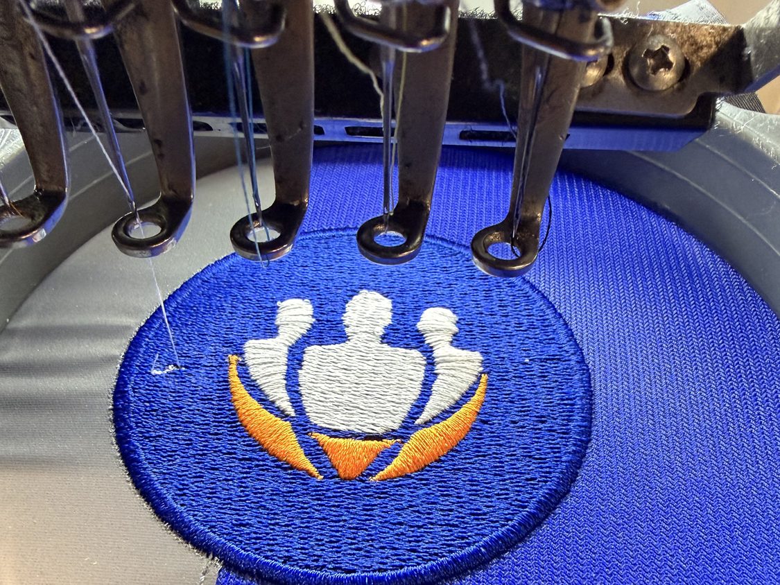 An embroidery machine stitching a blue and orange emblem into a blue fabric hoop