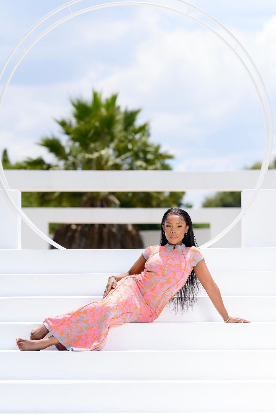 A woman reclining on white steps in a pink and orange patterned dress
