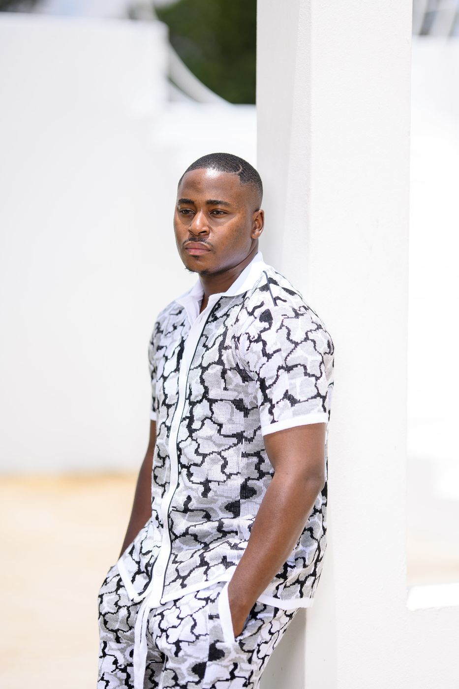 A man in a black and white patterned short-sleeve shirt leaning on a white wall