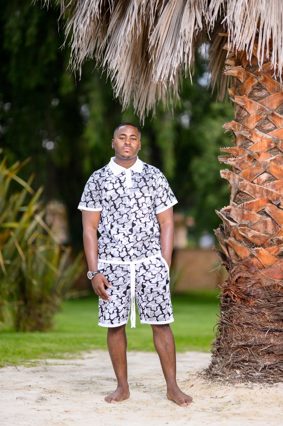 A man in a black and white patterned polo and shorts set under a palm tree