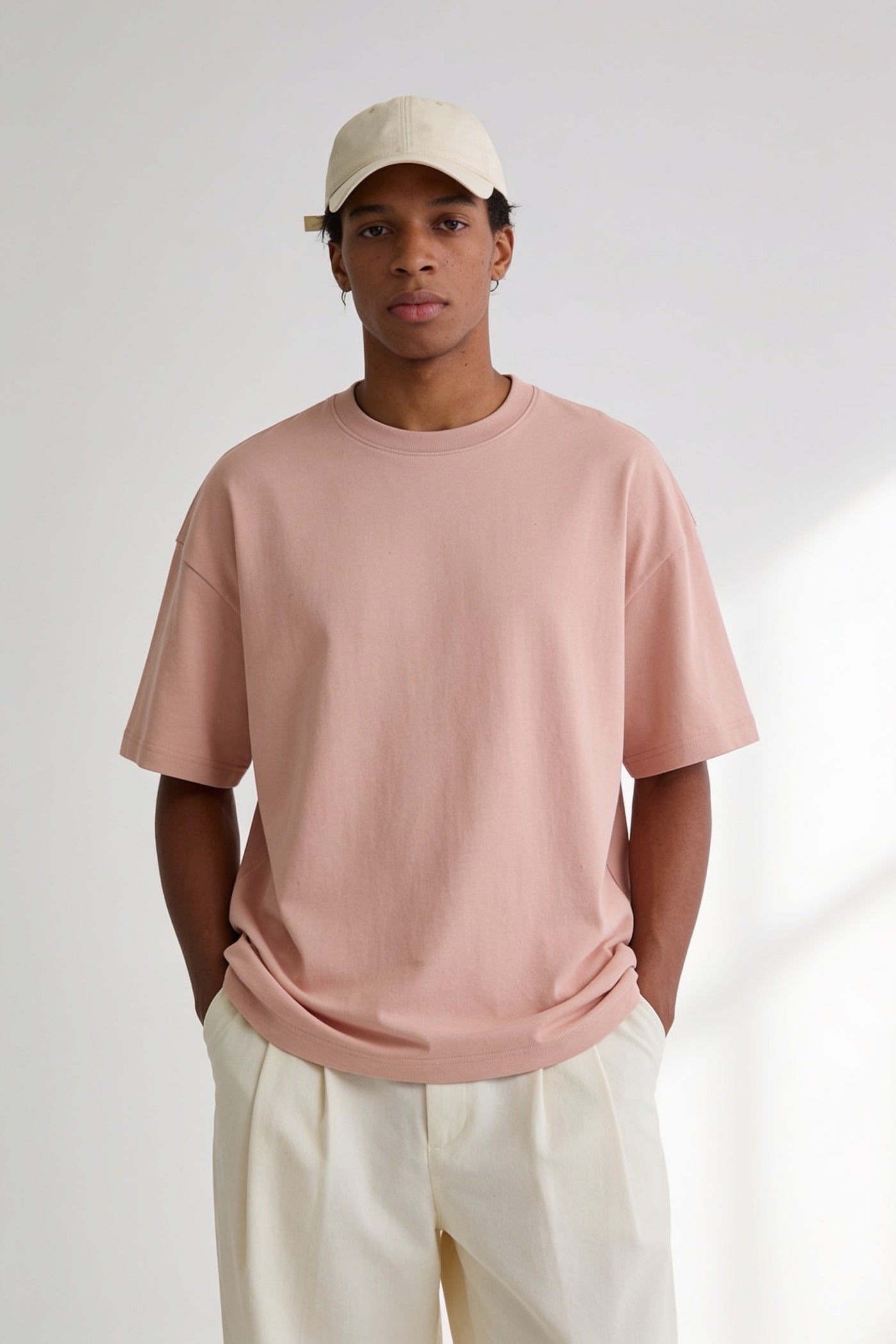 A young man in an oversized dusty pink t-shirt and a cream cap in a bright studio